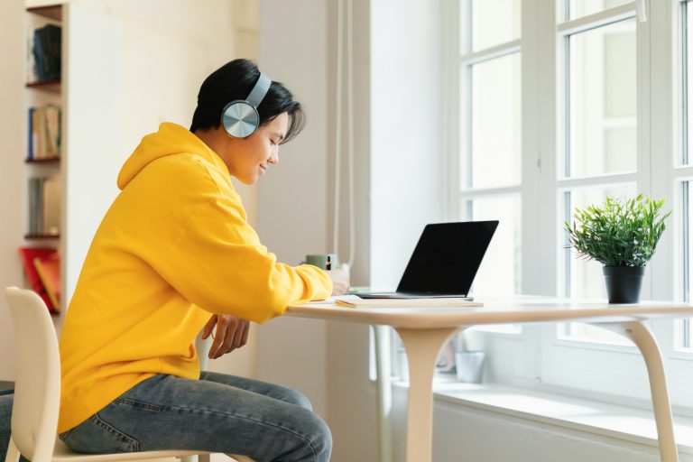 E-Learning. Asian Teenager Guy Sitting At Laptop Doing Homework Wearing Earphones And Taking Notes Sitting At Table At Home. Smiling Student Writing Essay Studying Online. Side View