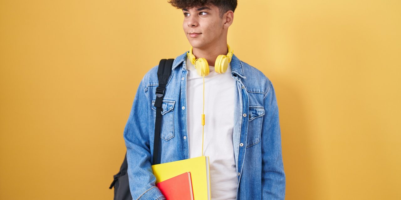 Hispanic teenager wearing student backpack and holding books smiling looking to the side and staring away thinking.