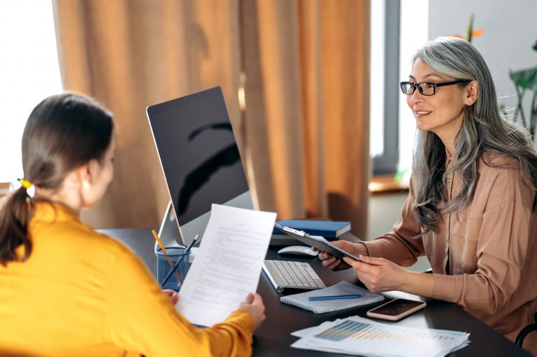 Confident intelligent mature gray-haired asian woman with glasses, business leader, HR manager, in modern office, talking to job seeker female, holding and leafing through resume, smiling friendly